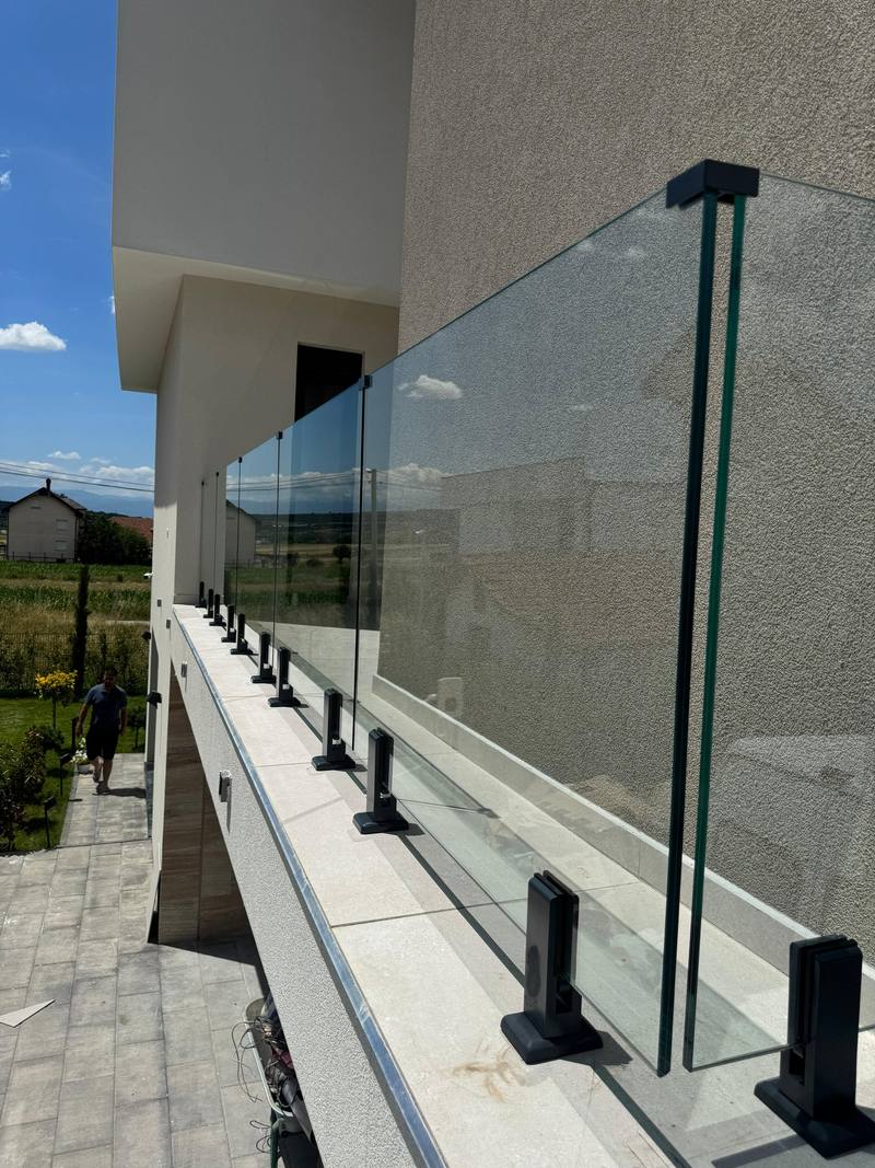 Parmak xhami në ballkon. Parmak me xham transparent përgjatë ballkonit të një shtëpie, me mbajtëse të zeza në pika. Fotoja 2 nga 2.